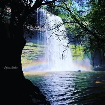 Place to See - Waterfall in Ghana with rainbow mist, cascading into a calm pool, and lush surrounded with greenery.